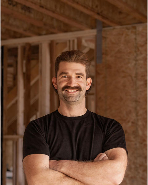 Denis Dume, smiling with arms crossed, standing in a framed house under construction