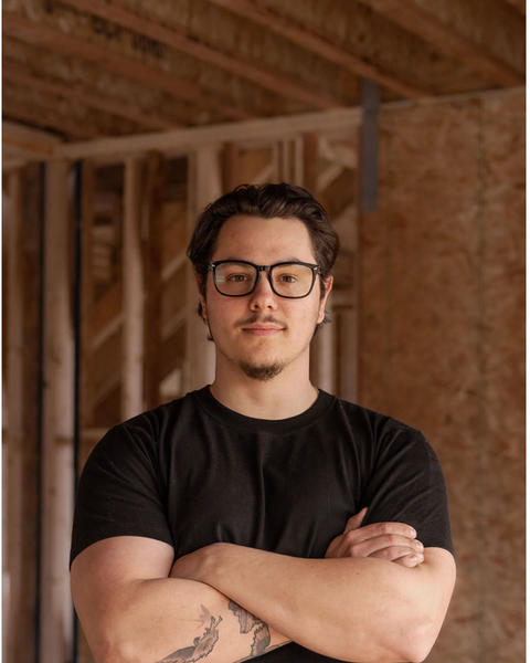 Maleky Colgiu, arms crossed, standing in a framed house under construction